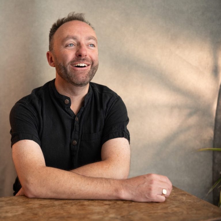 A photo of the vocal coach, Jamie Read, laughing towards the camera. He is wearing a black shirt and leaning on a wooden table